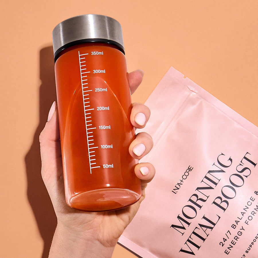 En hånd holder shakeren med appelsinfarget morgendrikk ved siden av posen med Morning Vital Boost