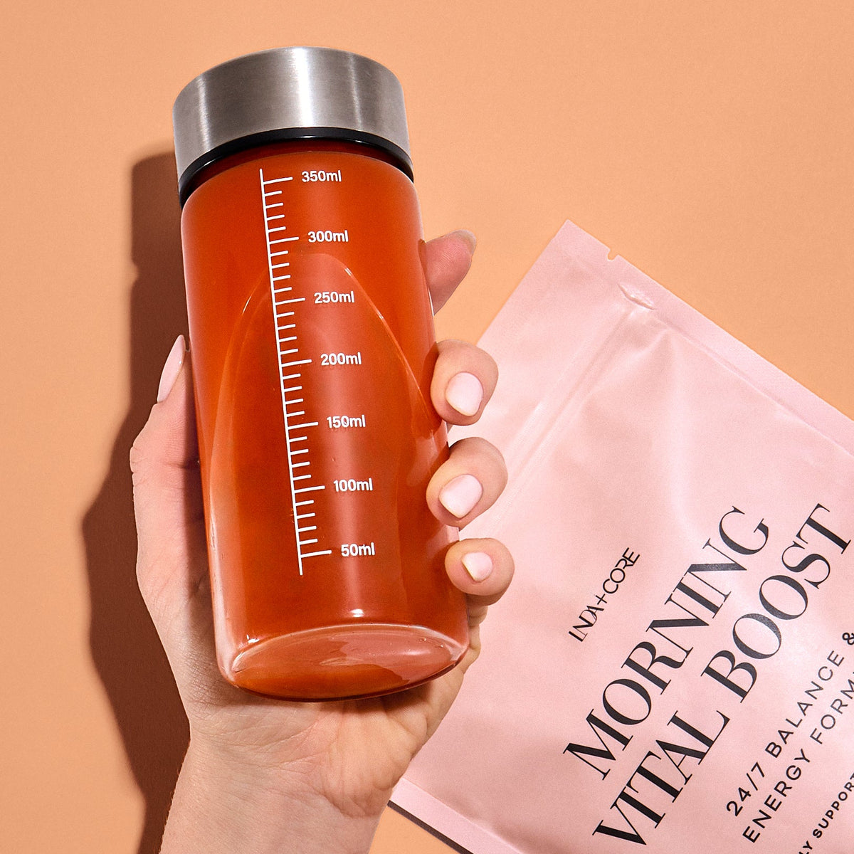 En hånd holder en shaker med appelsinfarget morgendrikk ved siden av posen med Morning Vital Boost